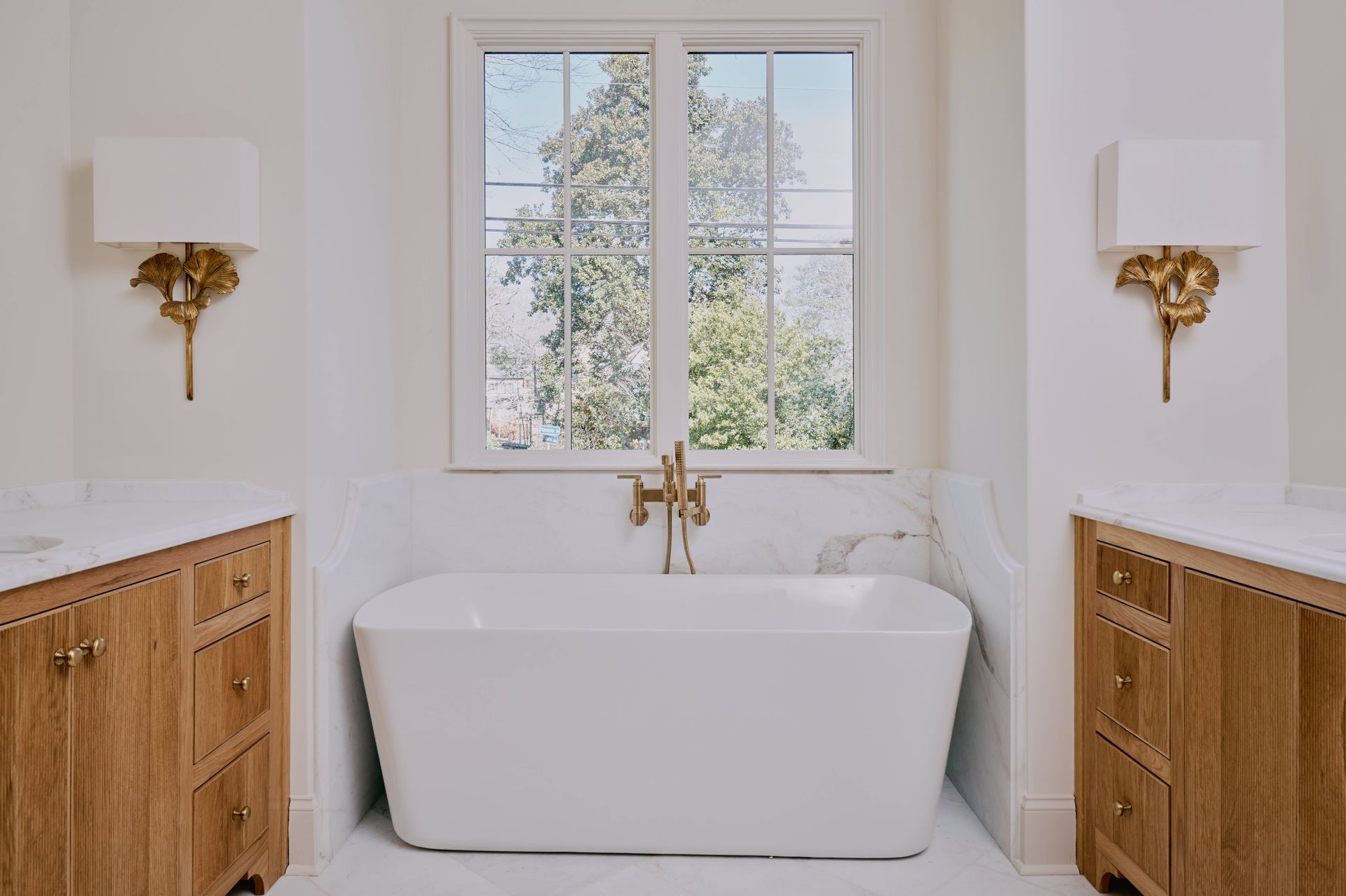 Bright bathroom with a white freestanding tub centered under a window and wood vanities on both sides.