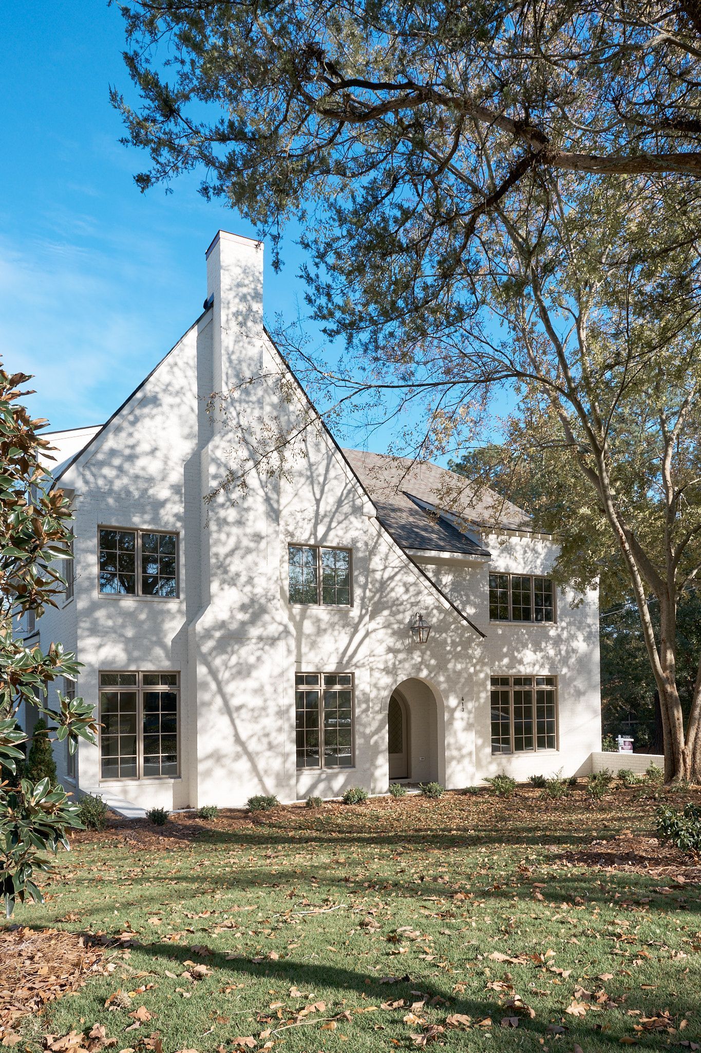 White house with steep roof and chimney under blue sky, framed by trees and autumn leaves