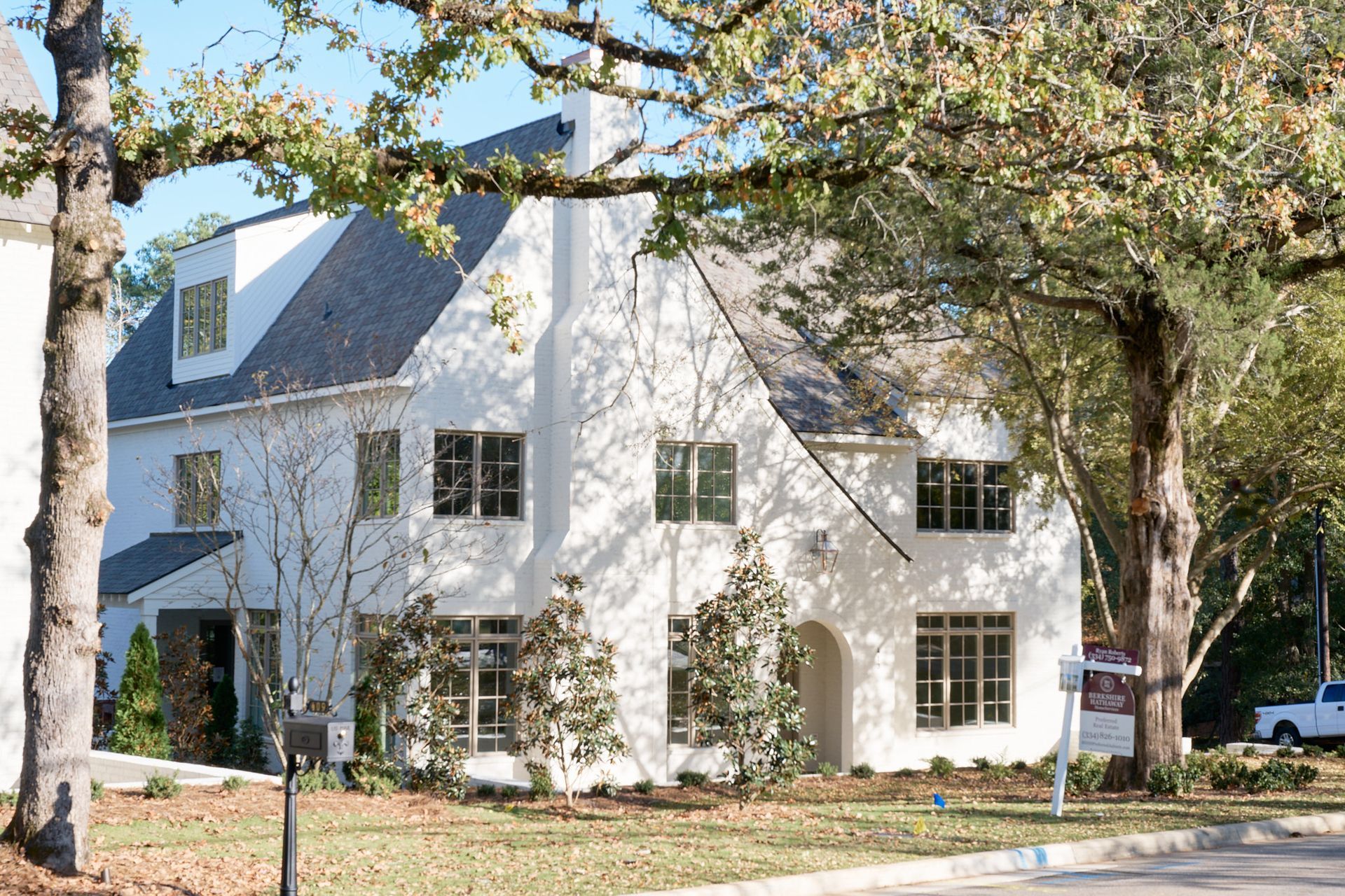 White two-story house with black roof and large windows, partially framed by trees along a quiet street