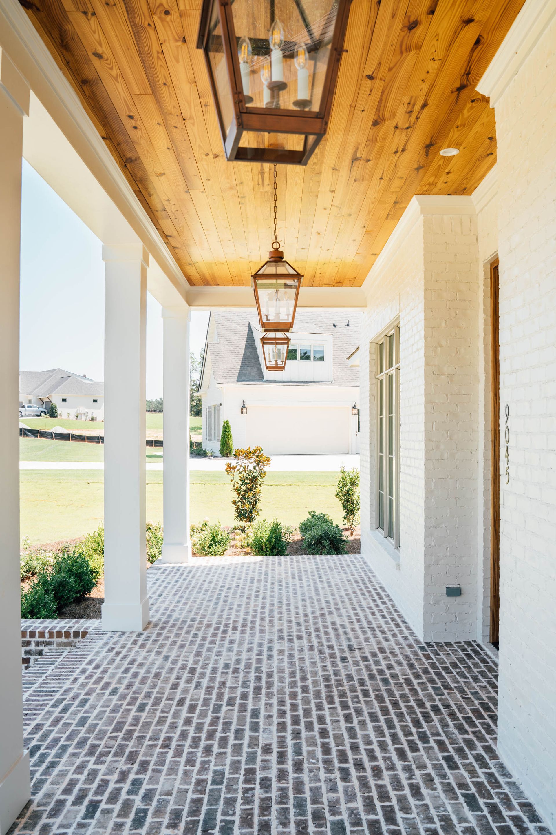 Covered brick porch with white columns, wood plank ceiling, and hanging lanterns leading to a bright yard