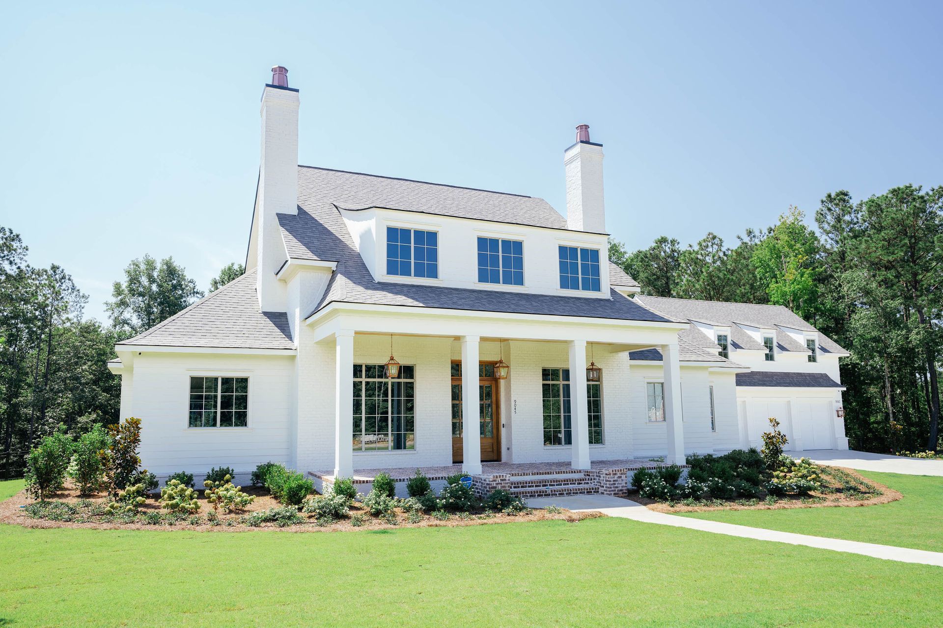 White two-story house with dormer windows, front porch, and a green lawn under a clear blue sky