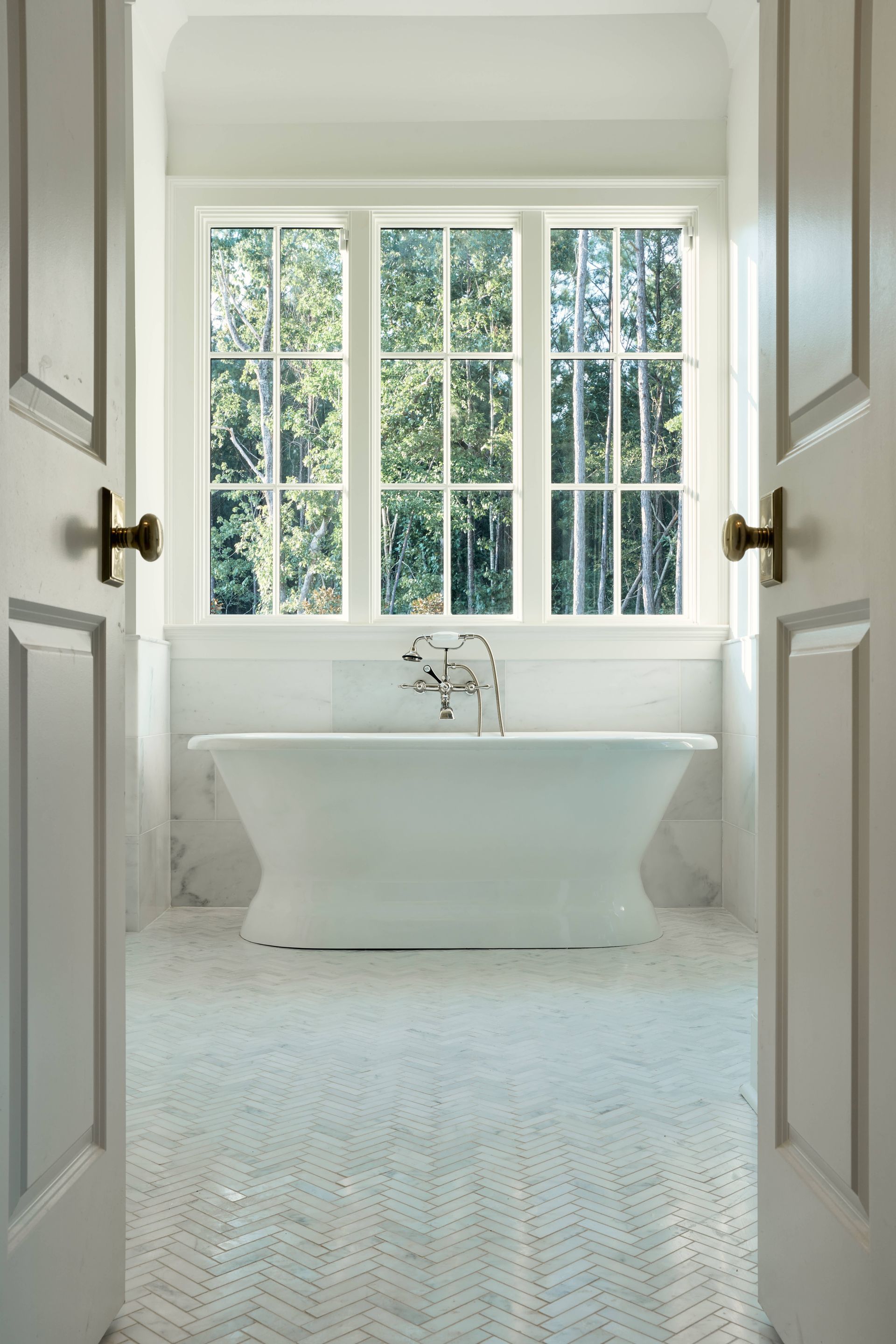 Bright white bathroom with a freestanding tub, large window, and open double doors.