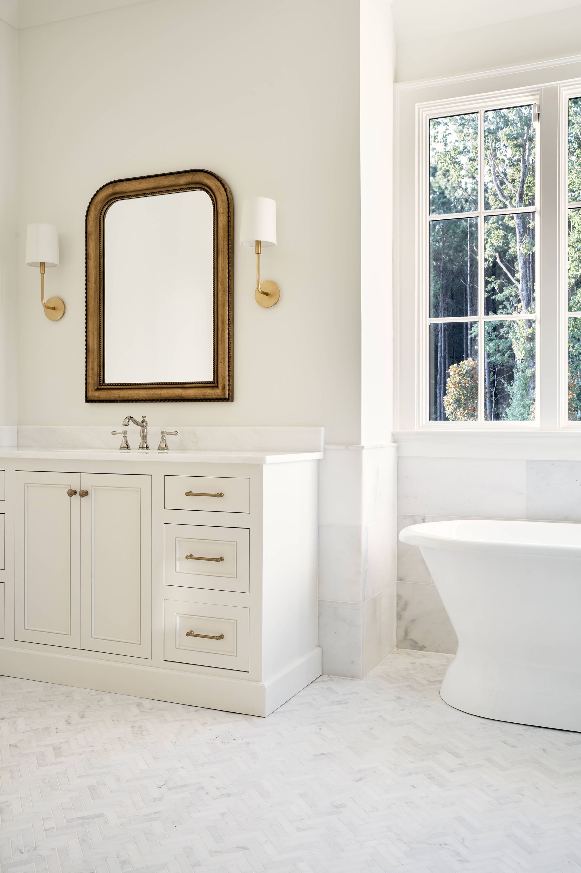 Bright bathroom with white vanity, gold-framed mirror, and window beside a freestanding tub.