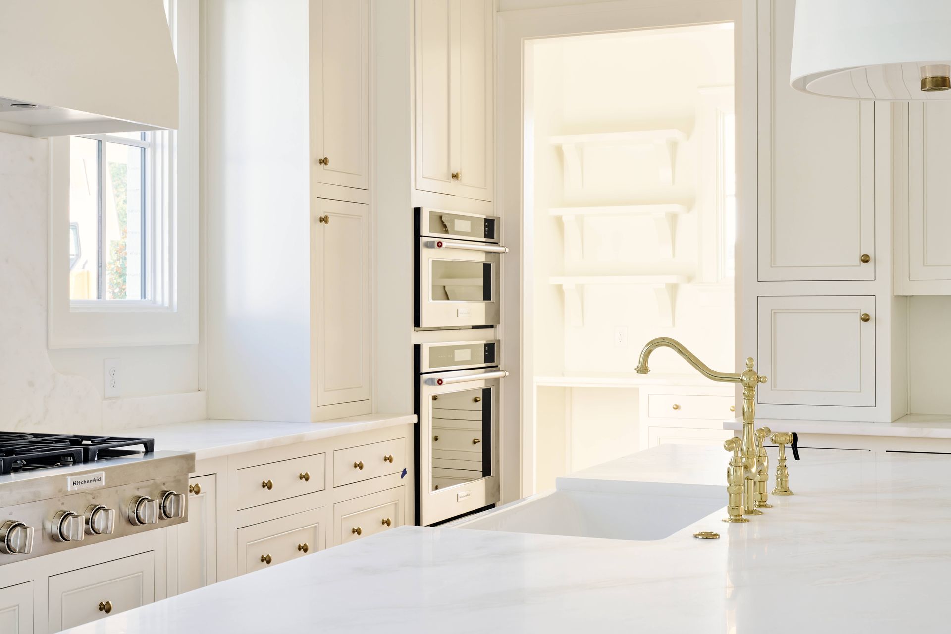 Bright white modern kitchen with island, gold faucet, built-in ovens, and large windows