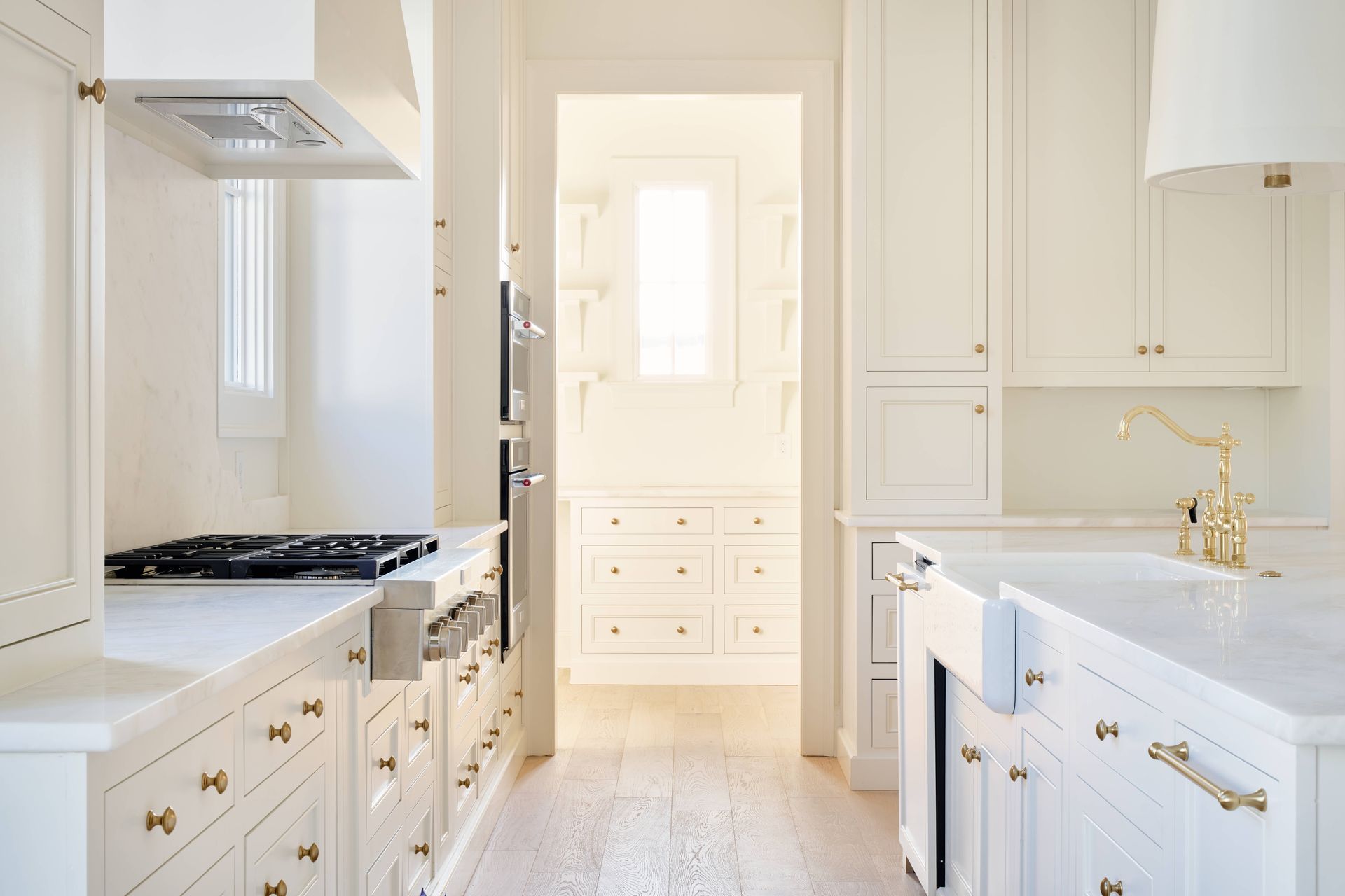 Bright white kitchen with gold accents, gas range, marble counters, and a doorway to a sunlit pantry.