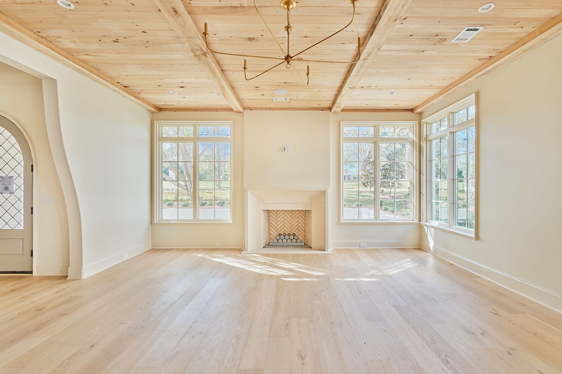 Bright empty living room with light wood floors, arched doorway, windows, and a small fireplace