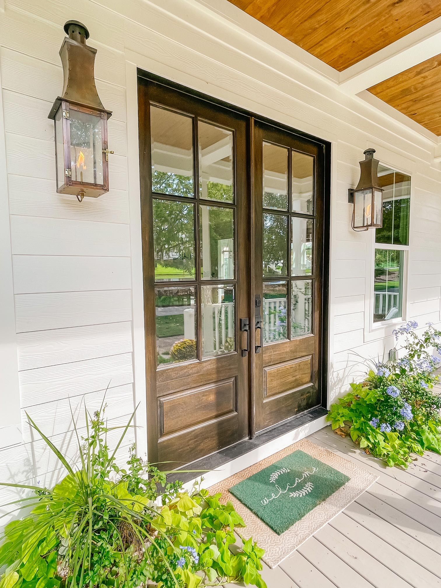 Front porch with wooden double doors, lanterns, potted plants, and a green doormat on white siding.