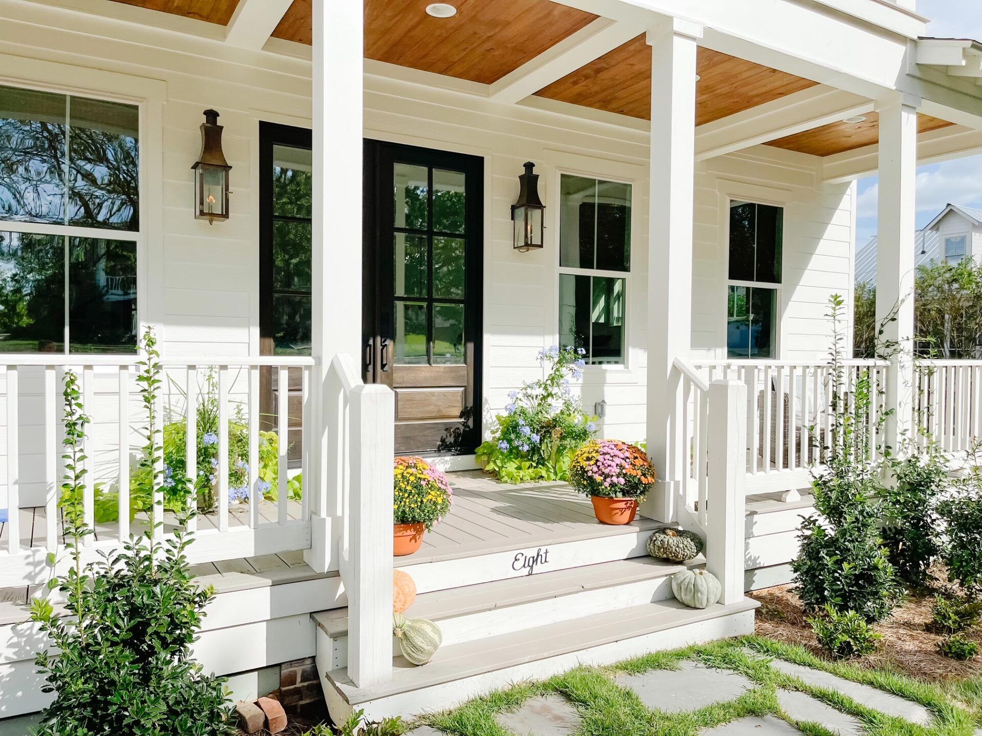 Front porch with white railings, black doors, potted flowers, and steps leading to a sunny house entrance.