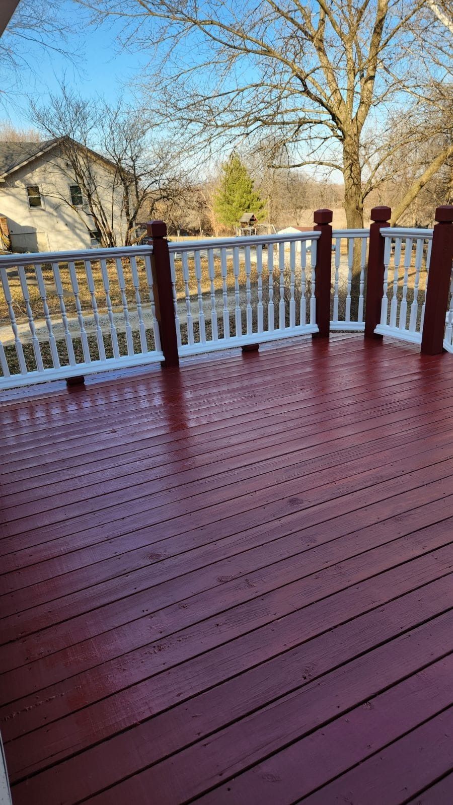 A wooden deck painted reddish-brown with a white railing, overlooking a yard with bare trees under a clear blue sky.