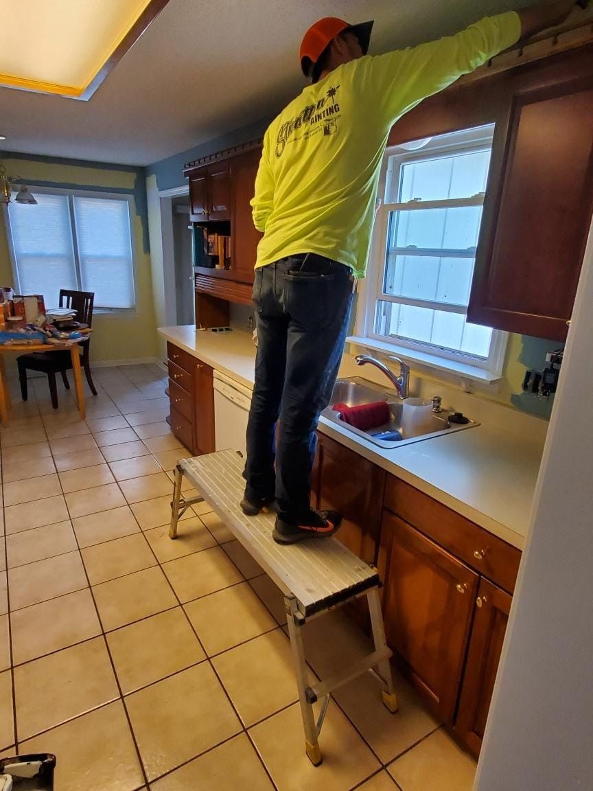 A person in a bright yellow shirt stands on a portable work platform to work on the ceiling above a kitchen sink.