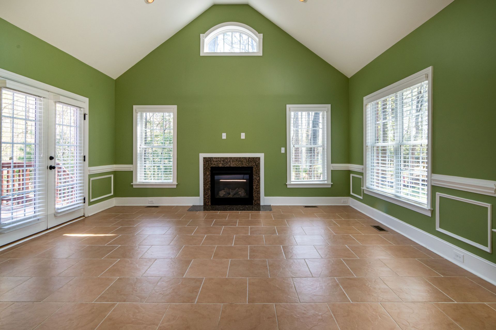 Empty living room with green walls, vaulted ceiling, large windows, and a central fireplace