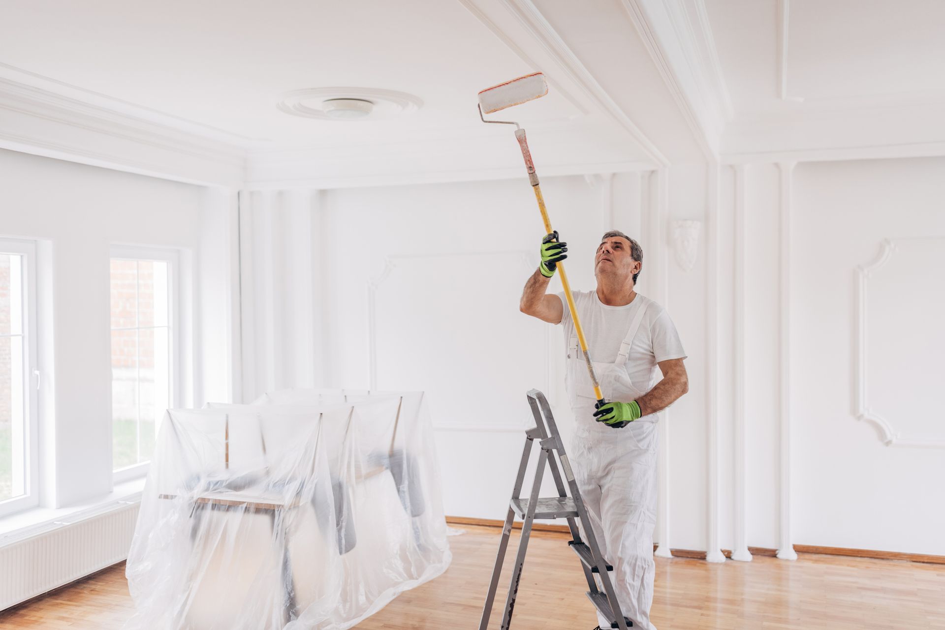 A painter standing on a ladder and performing interior surface painting on a ceiling.