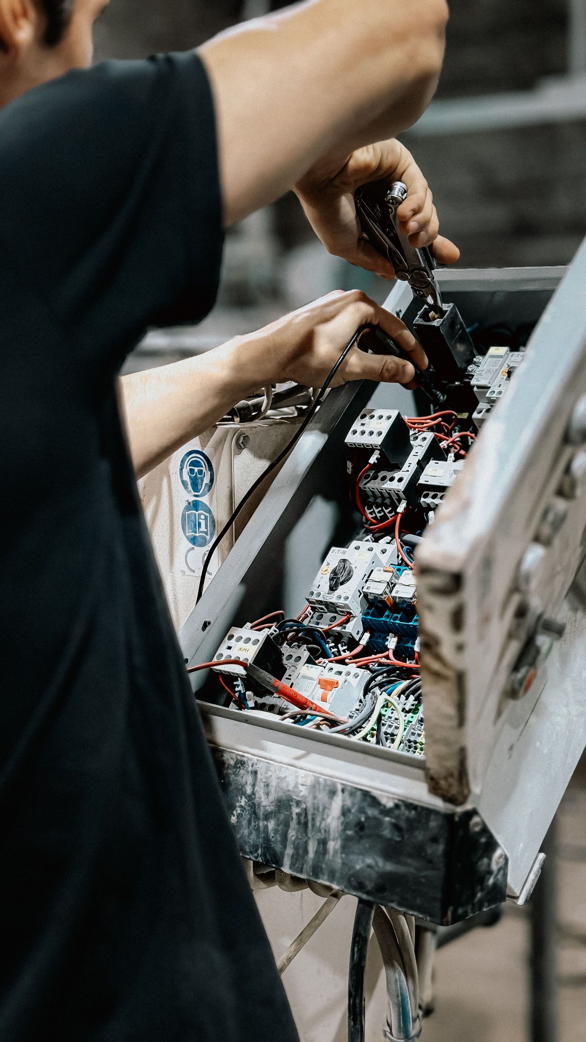Electrician testing wiring in an open electrical panel with a multimeter and screwdriver.
