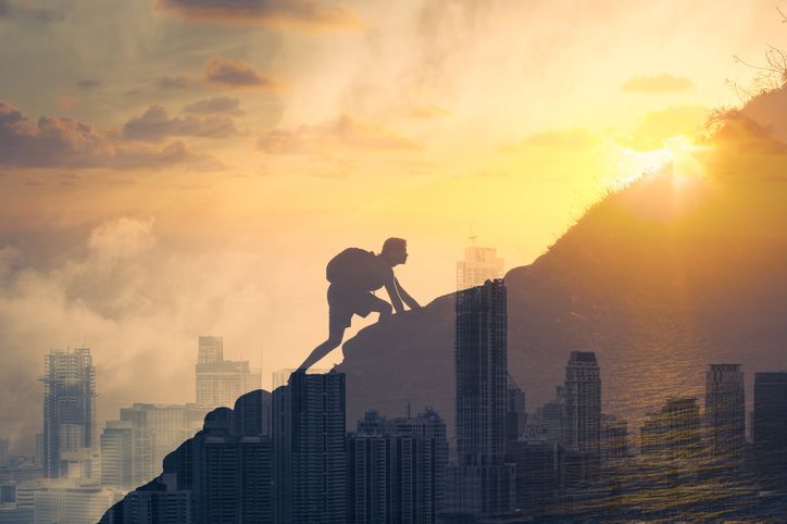 Image of man scaling mountain overlaid on an image of a big city with sun breaking through in the sky