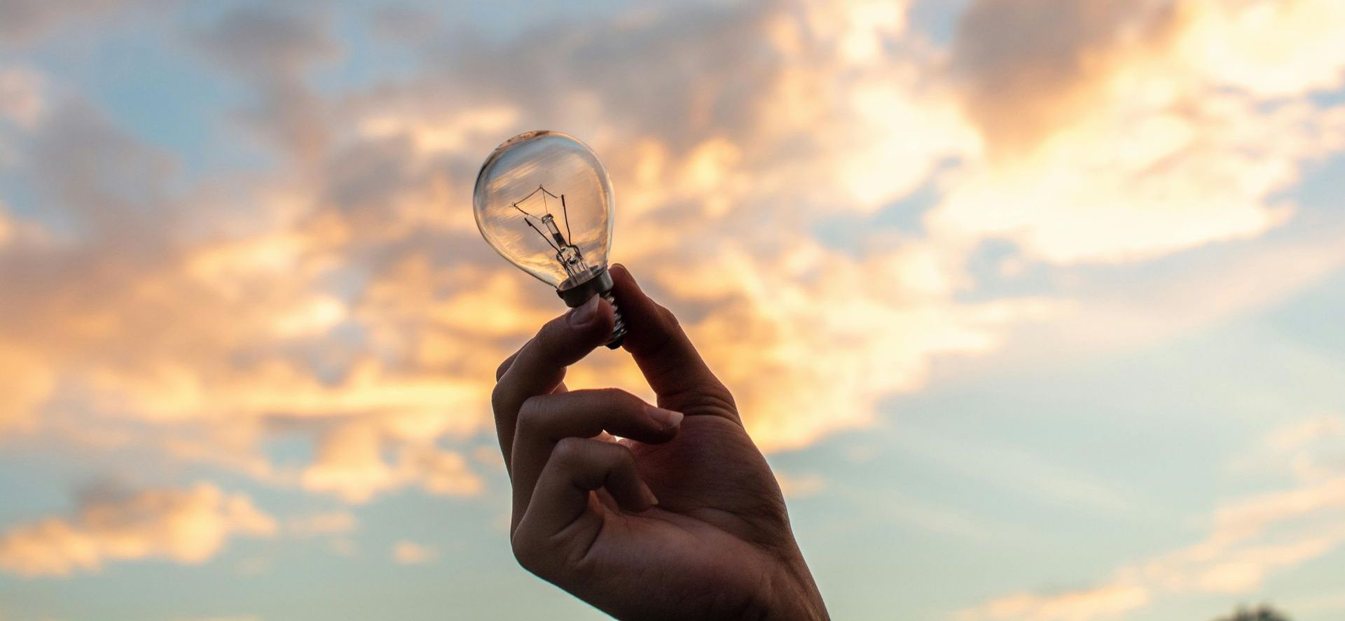 A person is holding a light bulb in their hand in front of a cloudy sky.