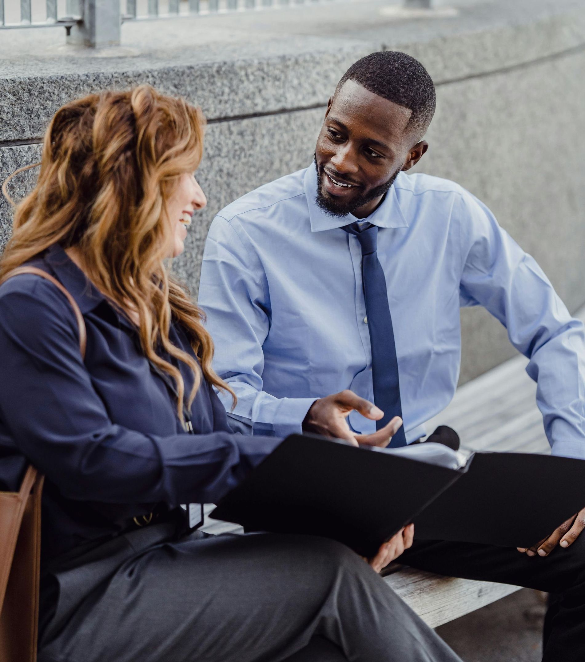 Businessman and businesswoman consulting over an open binder, seated outdoors on a bench.