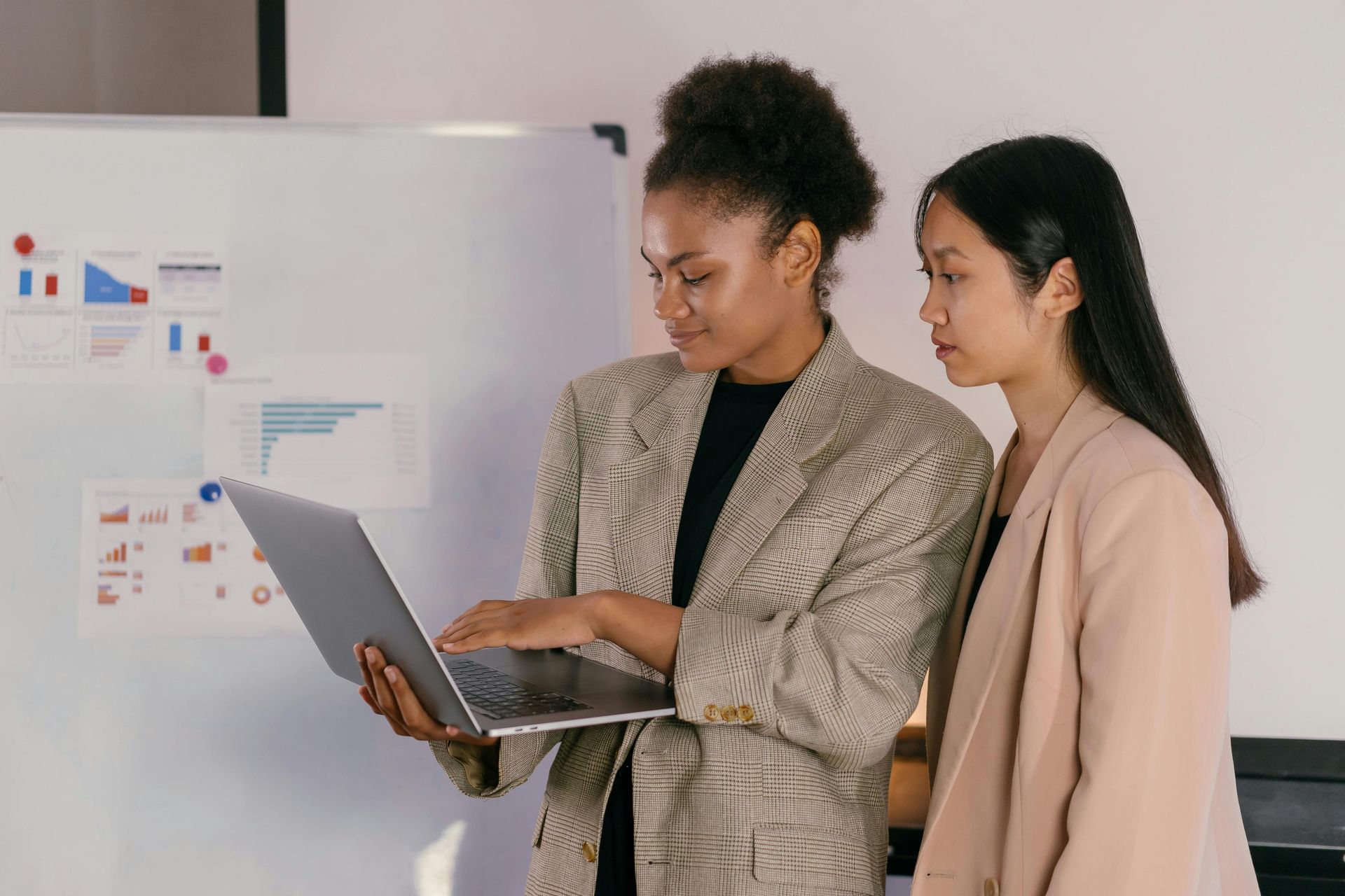Two women stand before a white board looking at information on a laptop