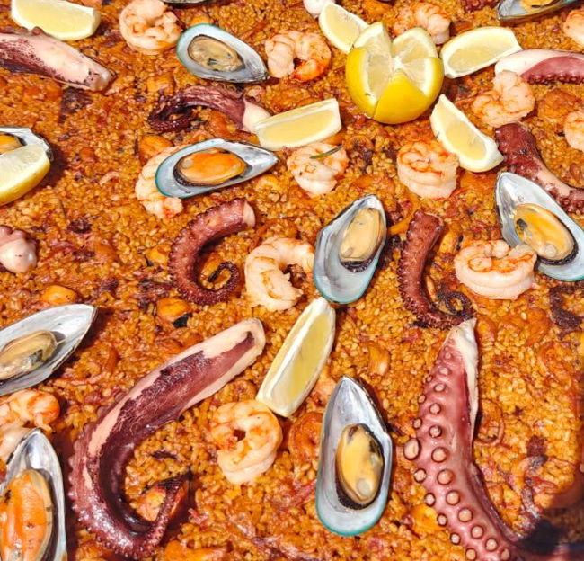 Paella with shrimp, mussels, and lemon wedges in a large, black pan.