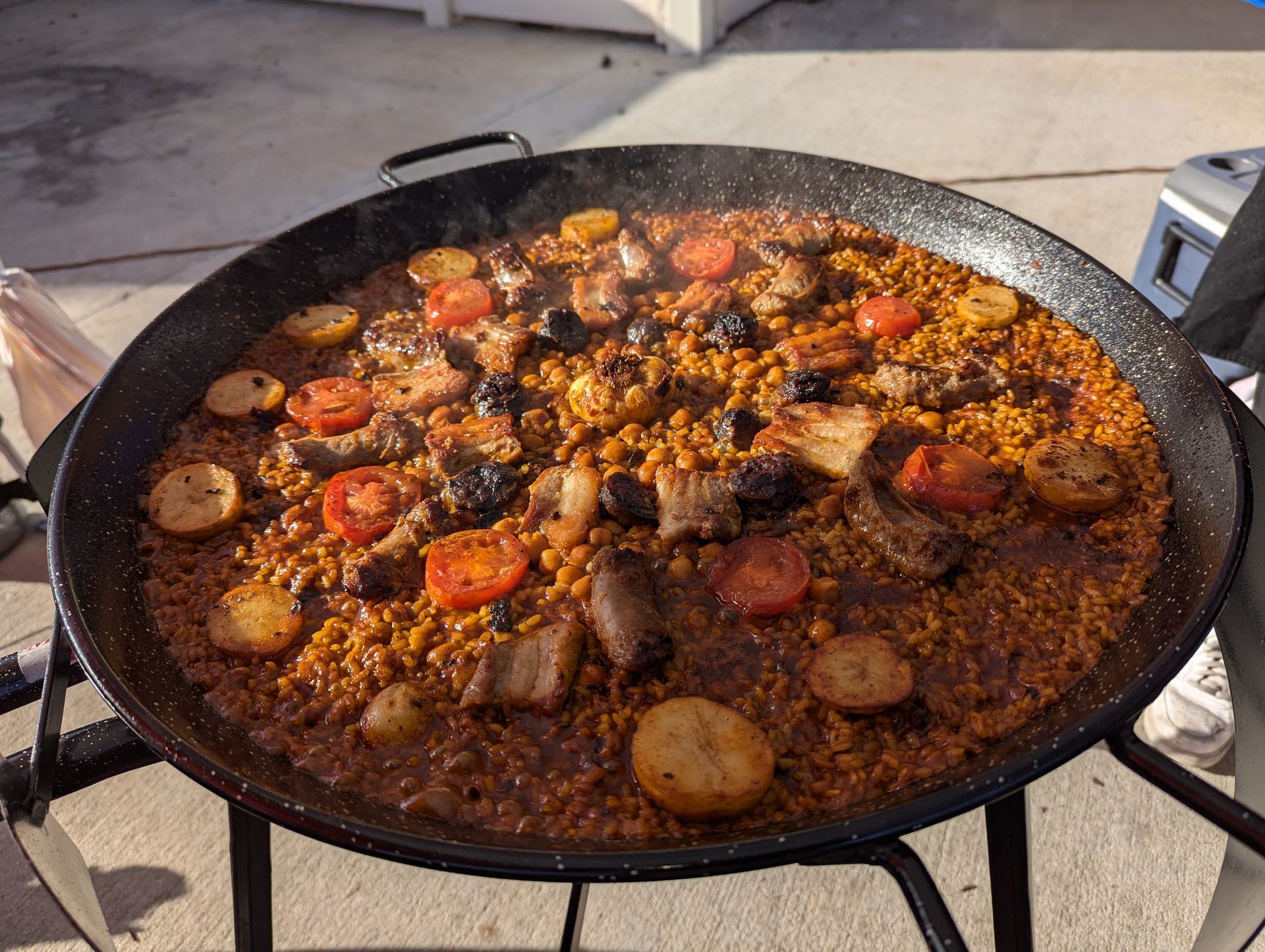 Paella cooked in a large pan on a propane burner, with rice, meat, potatoes, and tomatoes.