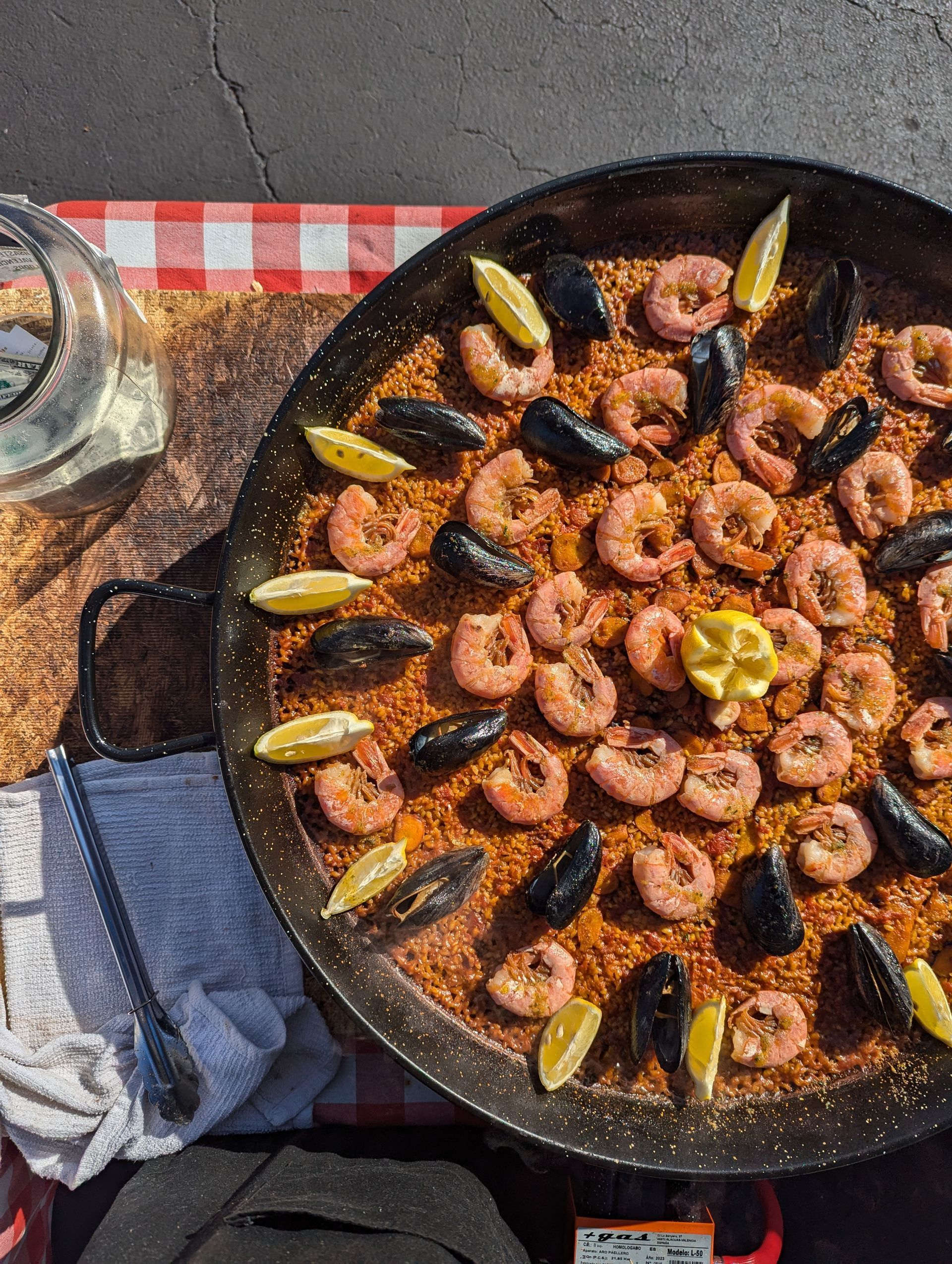 Paella with shrimp, mussels, and lemon wedges in a large pan on a red and white checkered tablecloth.