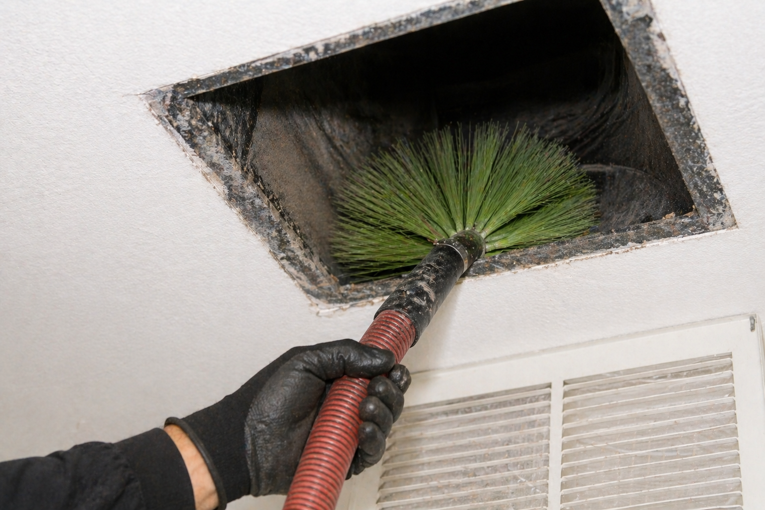 Gloved hand cleaning dust from an air vent with a red vacuum hose