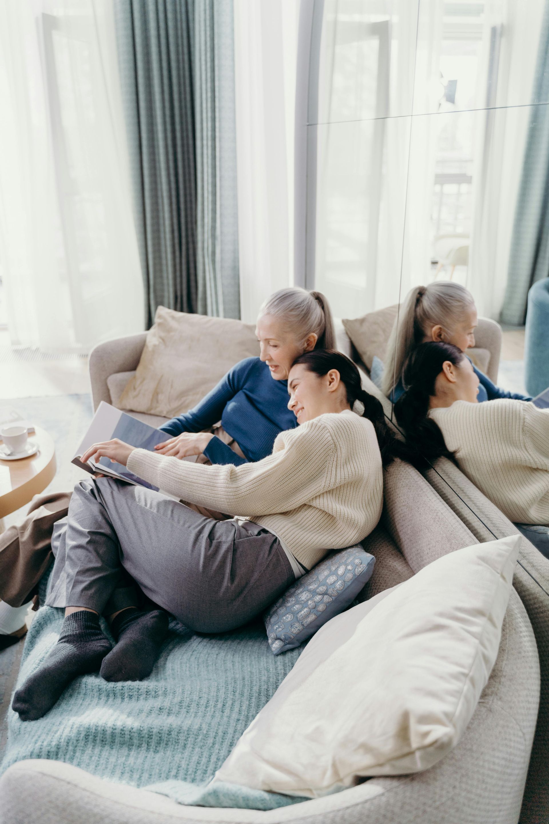 Two people lounging on a sofa with a laptop and tablet in a bright living room by the window