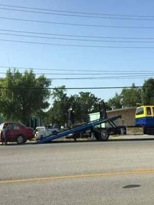 Tow truck hauling a blue vehicle on a flatbed beside a red car on a sunny street.