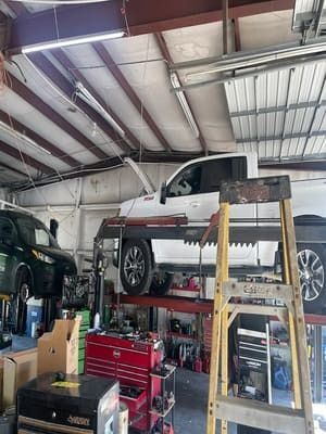 Auto shop interior with a white pickup on a lift, tools and equipment below, and a ladder in the foreground