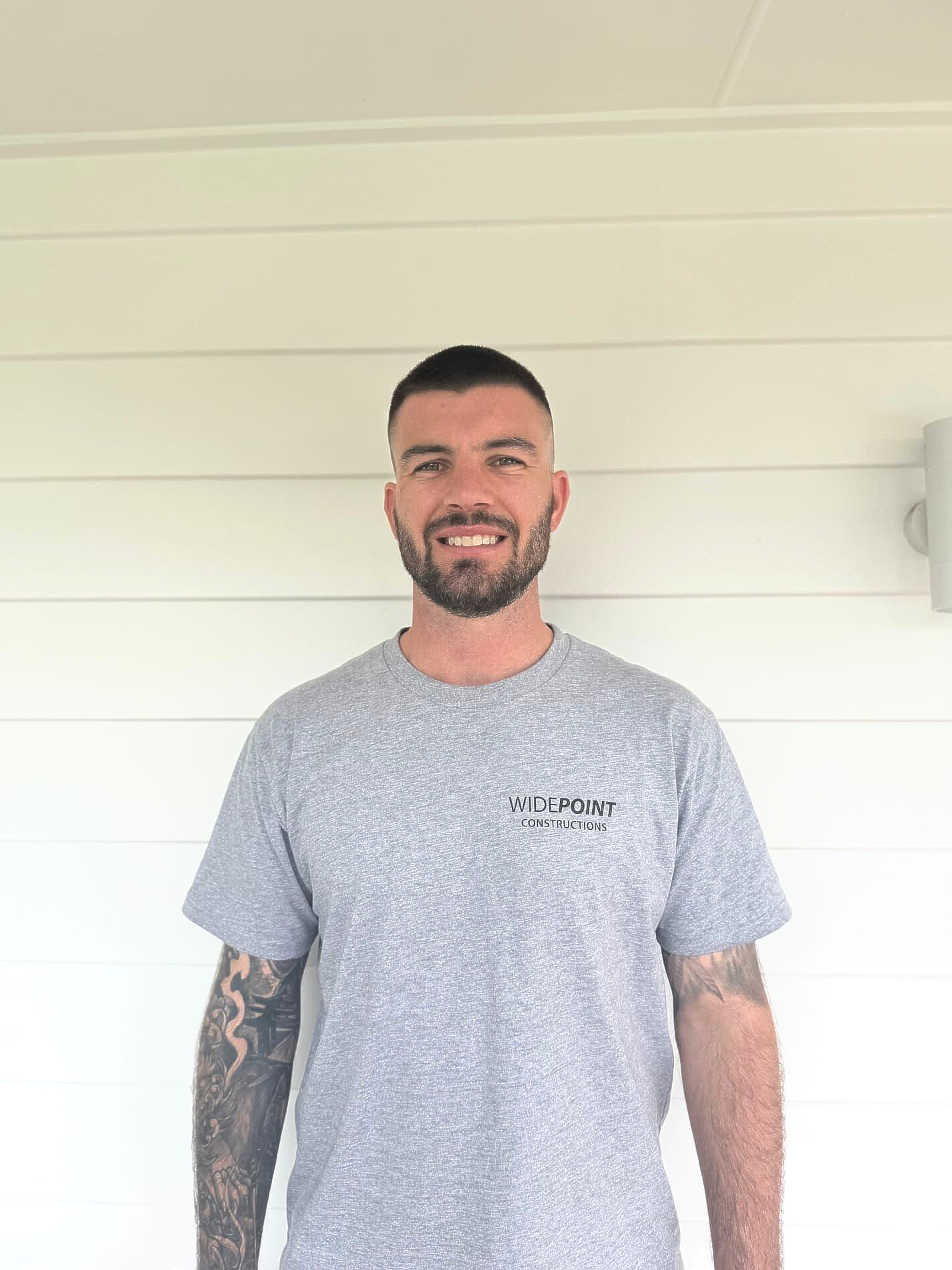 Man in work clothes poses on a porch, smiling. He wears a black shirt, tan shorts, and a tool belt.