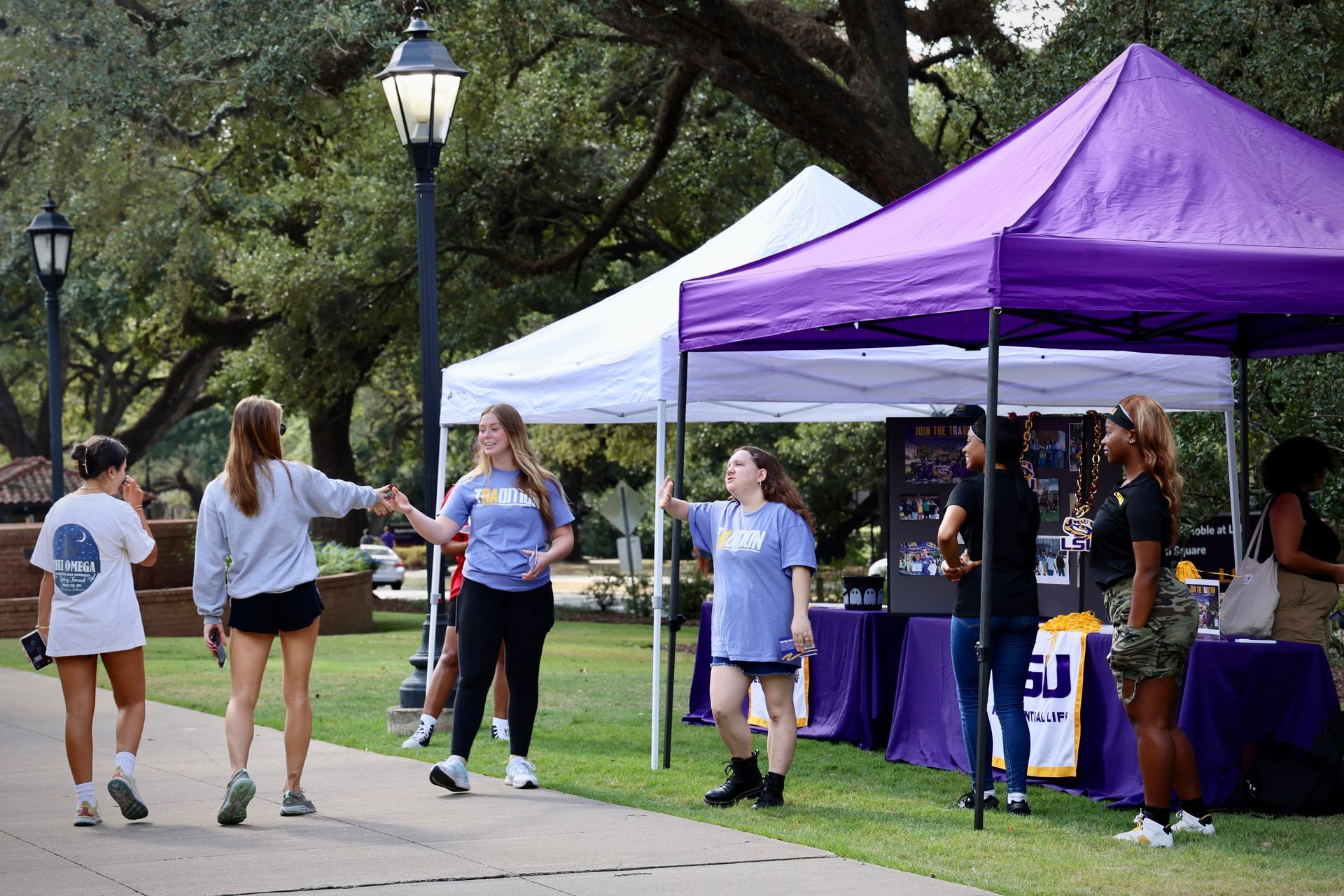People walking past a purple event tent in a park with trees and a lamppost