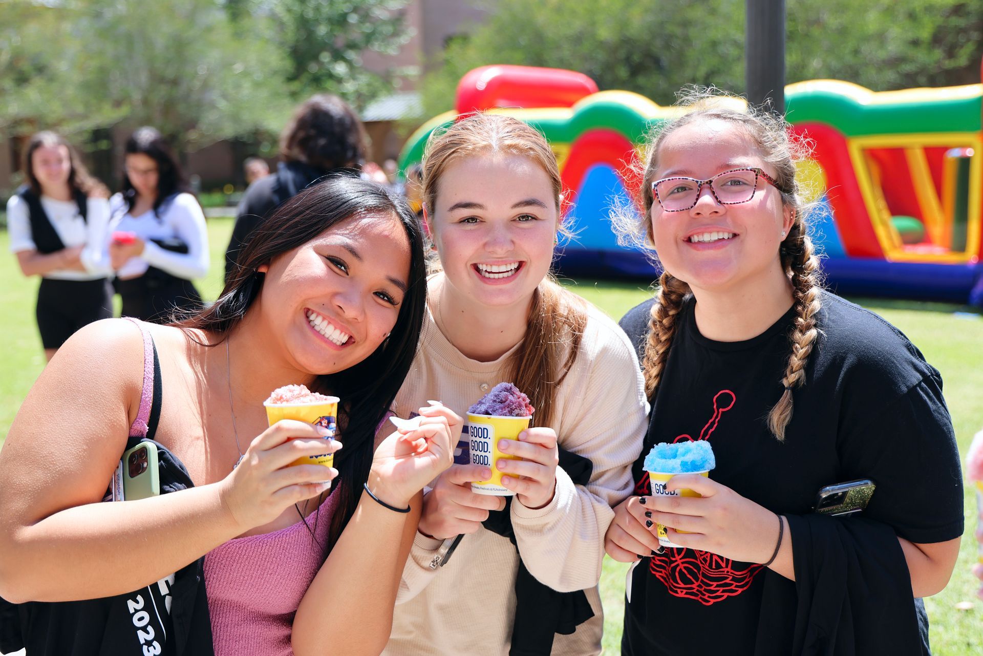 Three smiling friends holding ice cream cups at a colorful outdoor event with an inflatable bounce house.