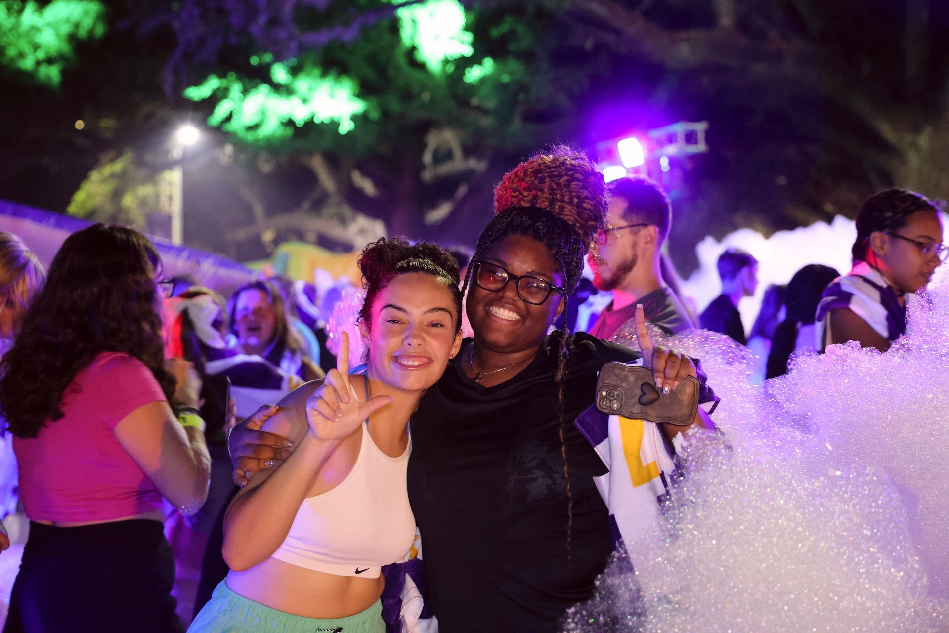 Two people smiling and posing at a lively night party with colorful lights and foam bubbles