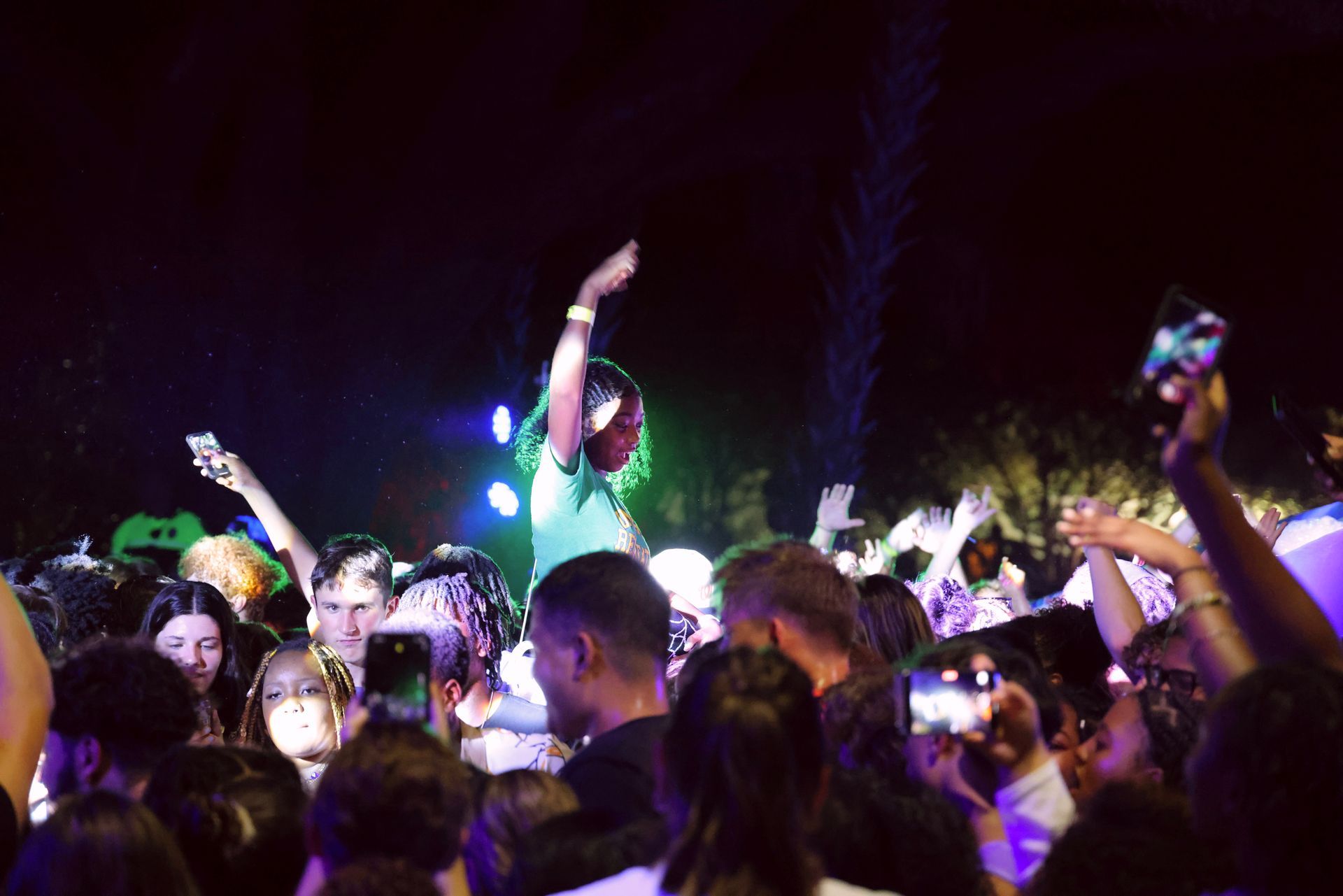 Crowd at a nighttime concert, with colorful stage lights and people raising their hands.