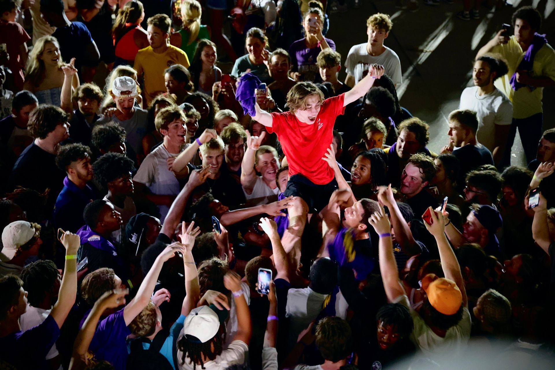 Crowd cheering as a person in a red shirt is crowd-surfed overhead at a concert