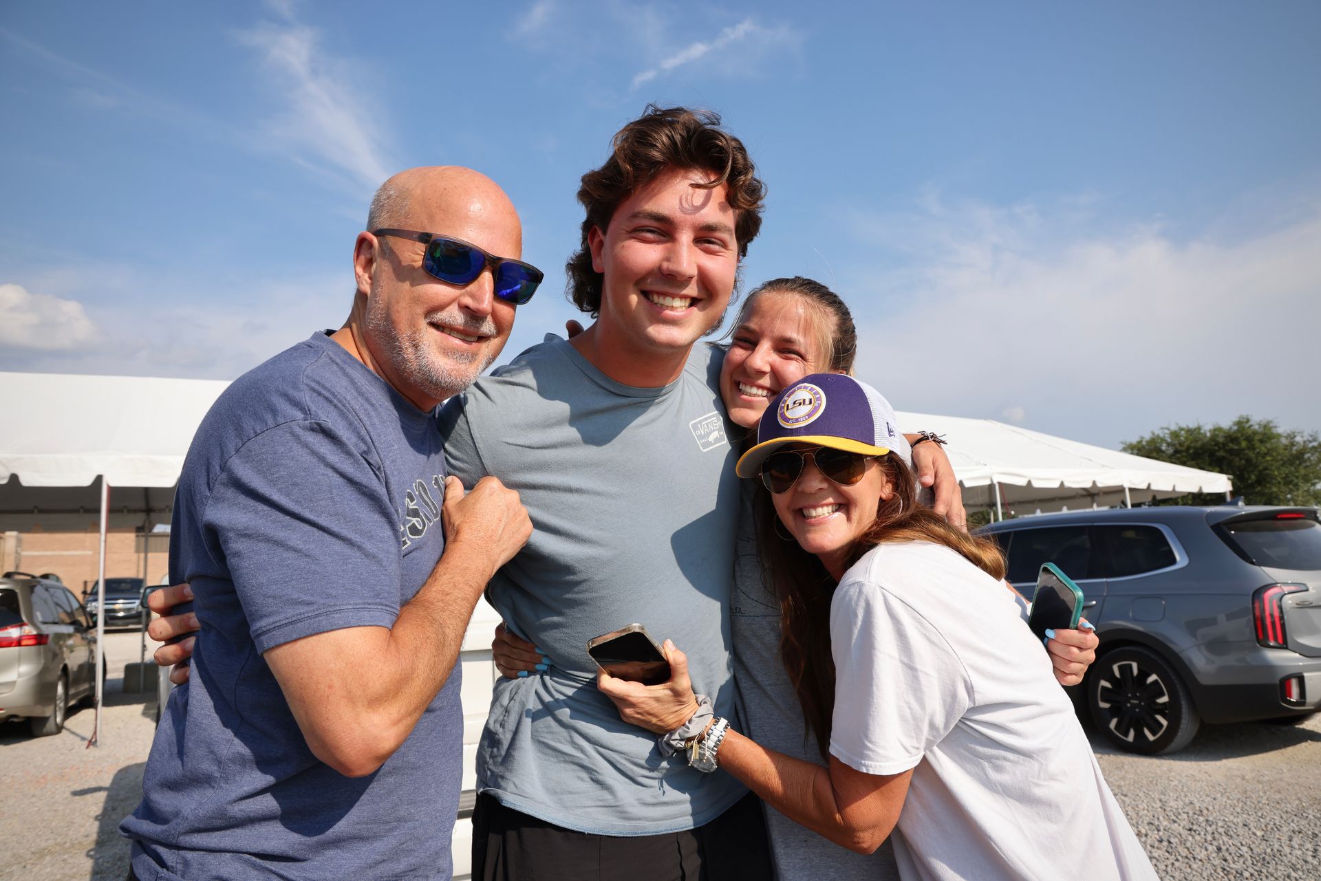 Three smiling people pose outdoors near parked cars under a sunny sky.