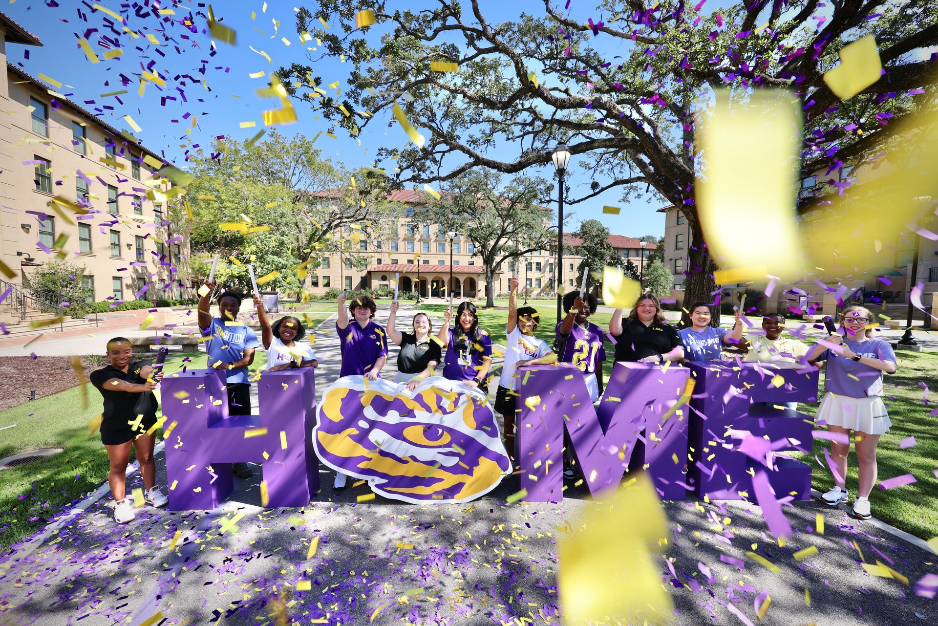 Group posing with purple “HOM” letters and confetti in a sunny campus courtyard