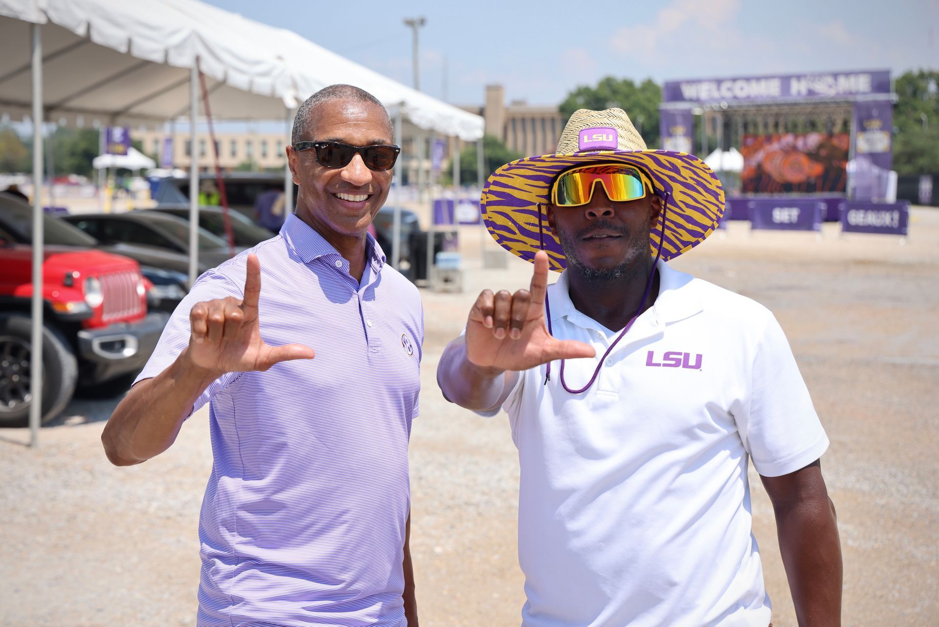 Two men smiling and posing with hand signs at an outdoor event with tents and cars behind them