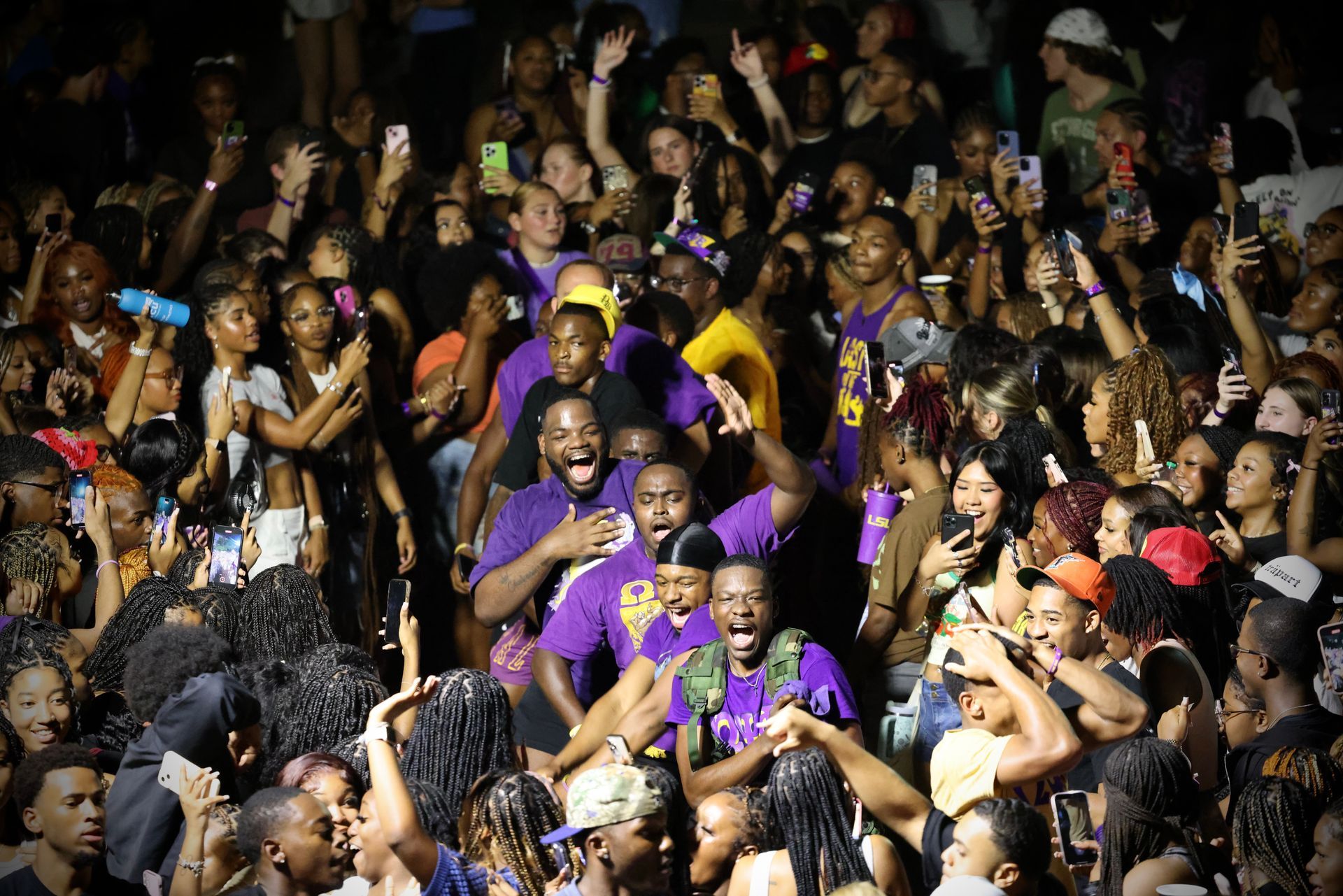 Crowd cheering at a concert, many people raising hands and phones under bright lights