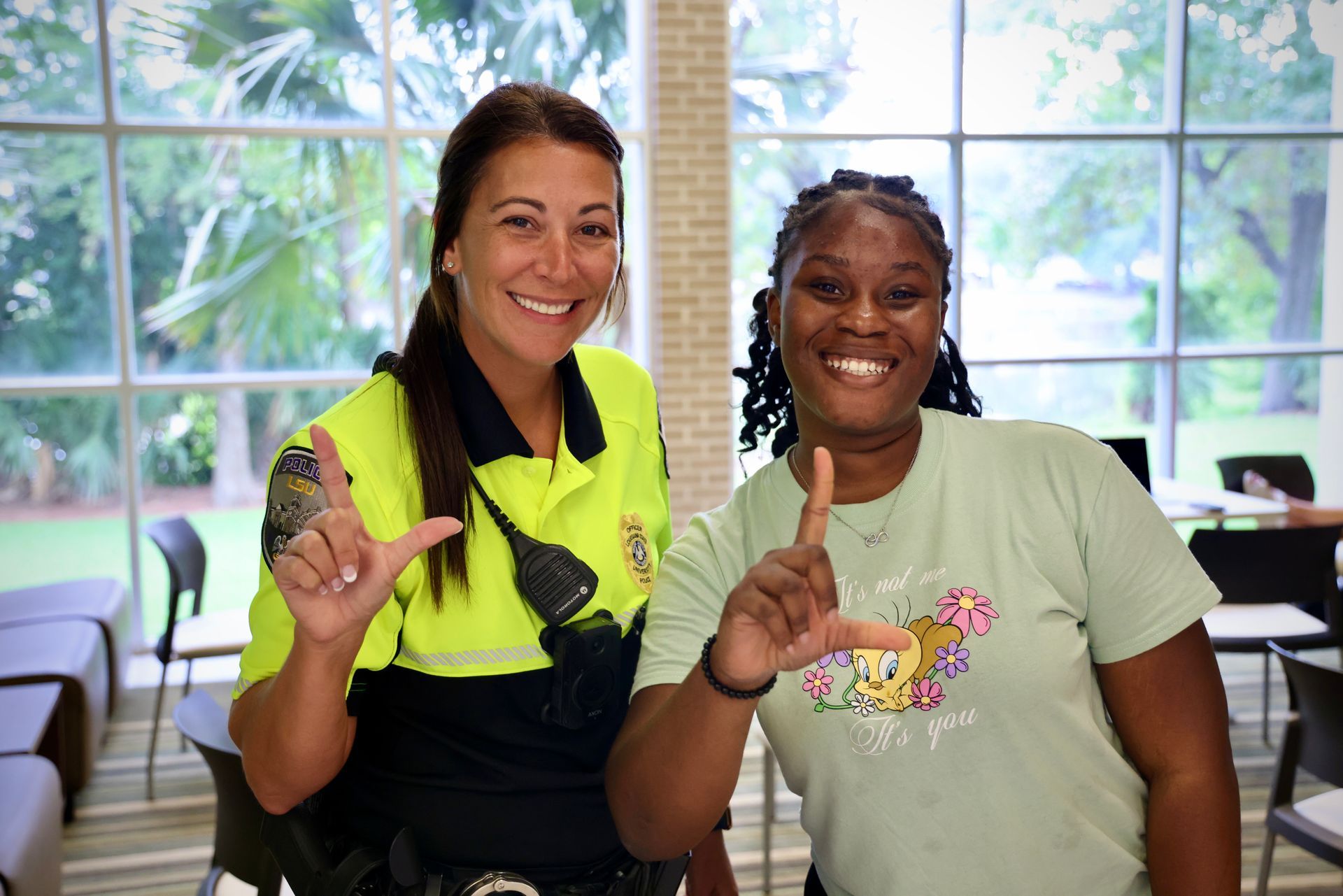 Two smiling people posing indoors, each holding up one finger, with large windows behind them.