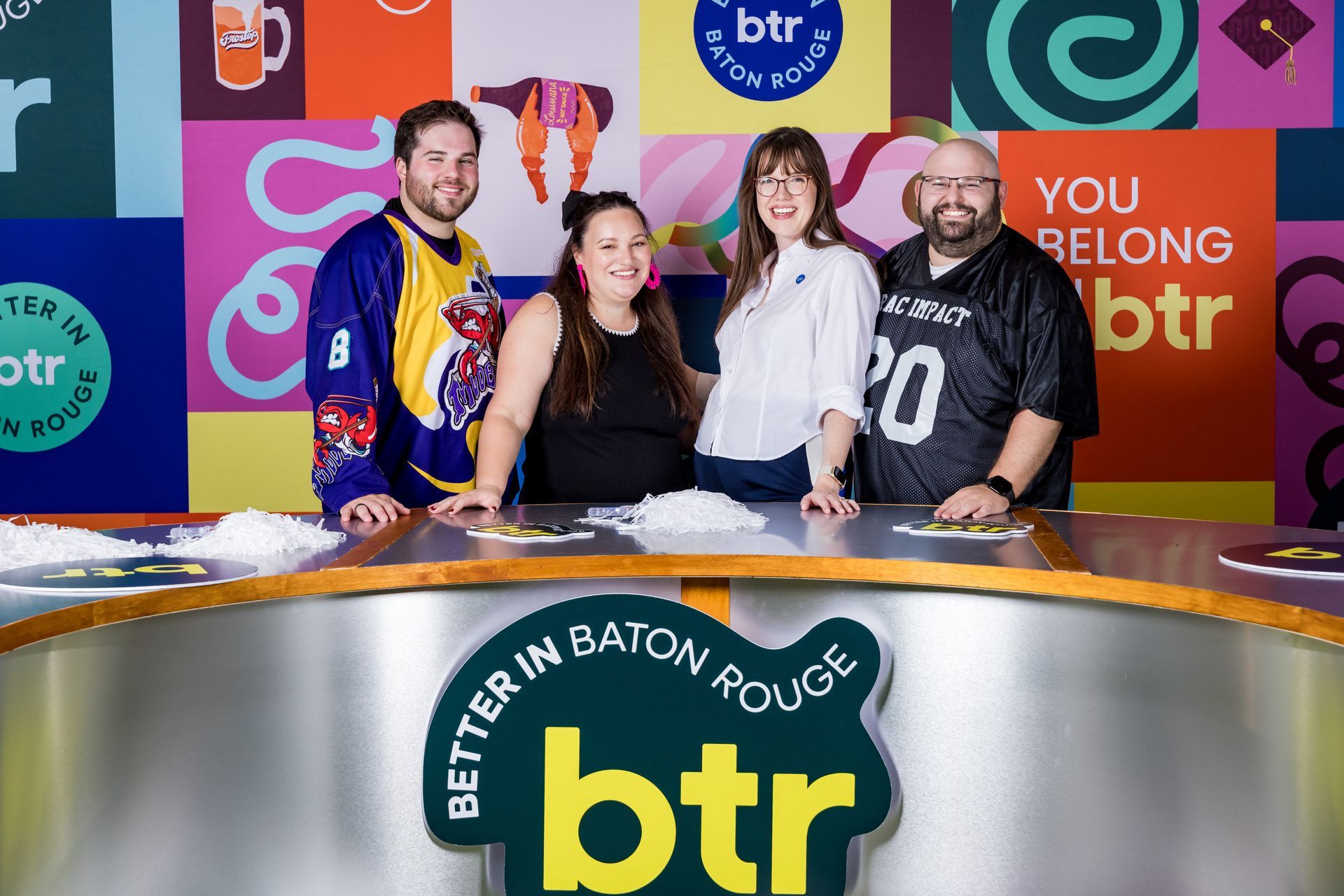 Four smiling people pose behind a BTR news desk in a colorful studio.