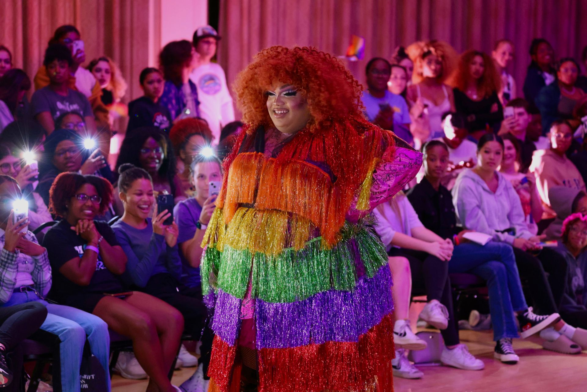Colorful performer posing on a runway in front of a seated audience under purple lighting