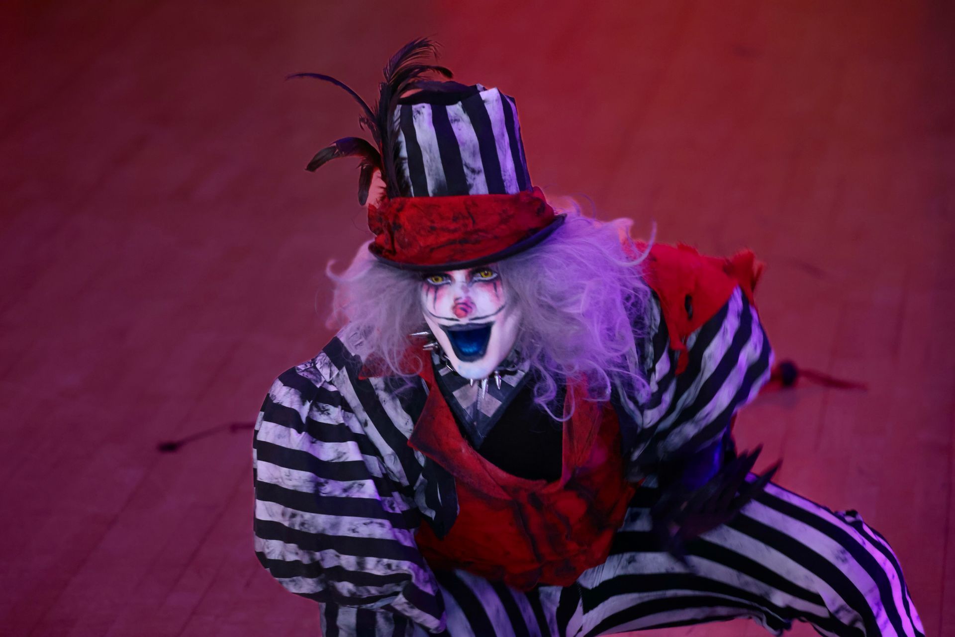 Clown in striped costume and top hat crouches under magenta lighting, looking downward