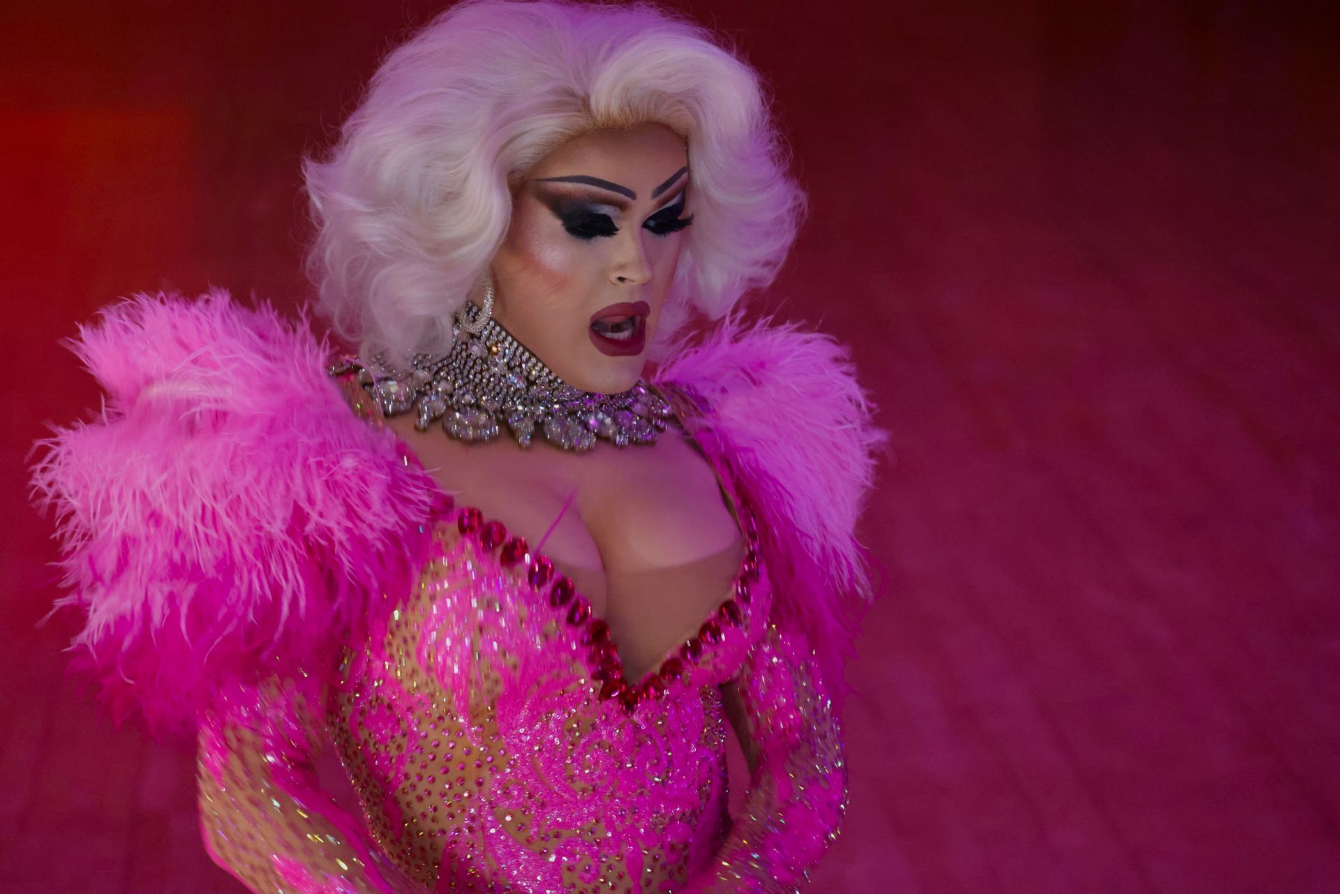 Person in a bright pink feathered outfit with platinum hair and dramatic makeup on a stage-like floor