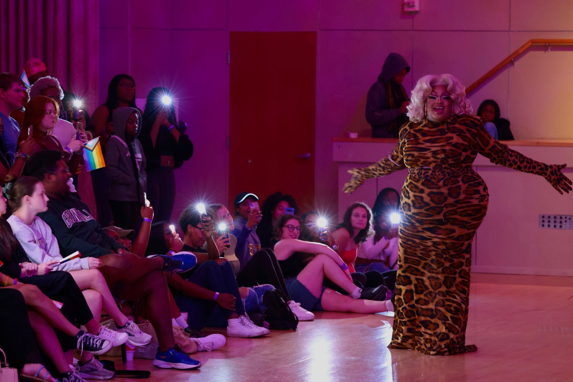 Drag performer in leopard-print gown posing onstage before seated audience holding up phone lights