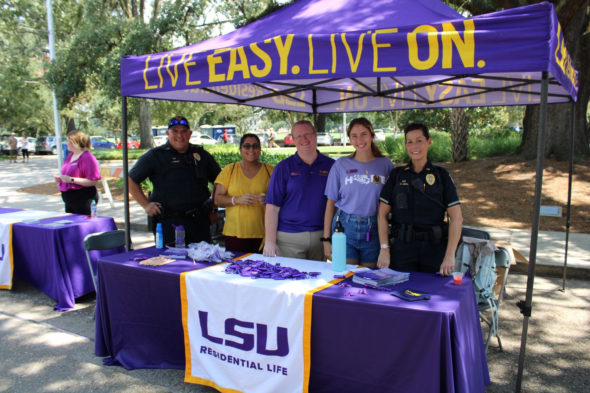 LSU Tailgating table under purple canopy with six people and “LIVE EASY. LIVE ON.” banner