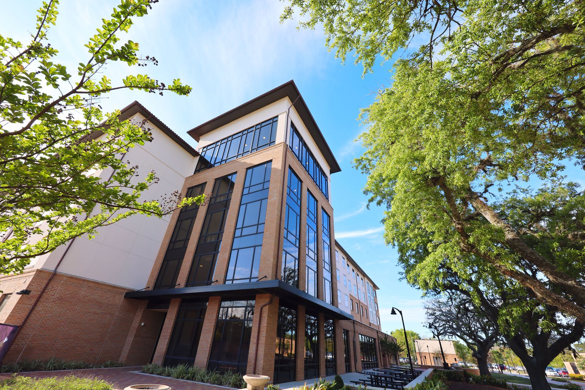 Modern brick-and-glass building with tall windows, framed by leafy trees under a blue sky.