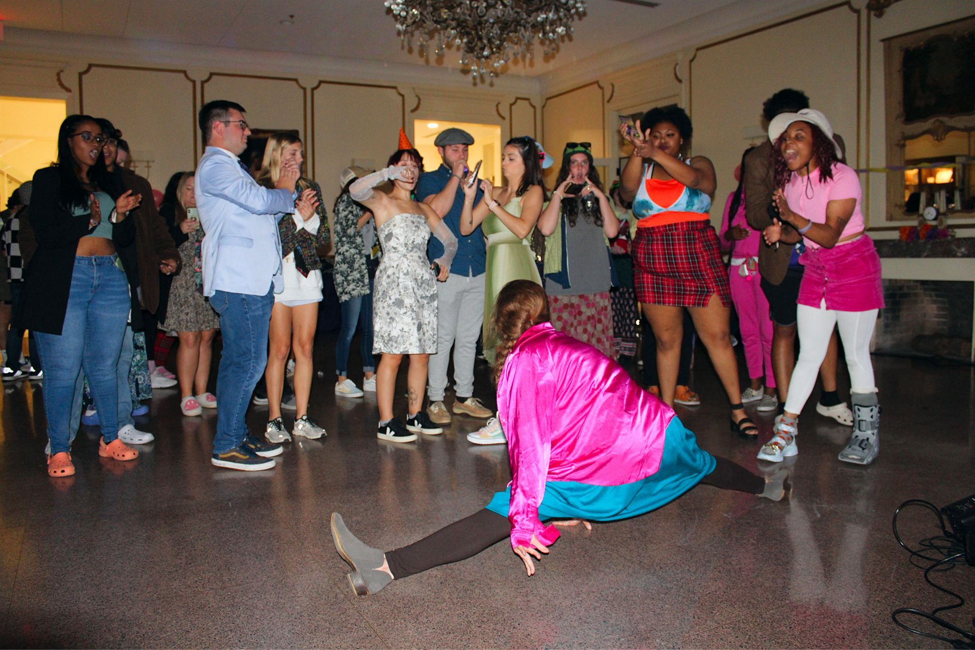 Person in bright pink and blue outfit dances on a ballroom floor while a crowd watches and cheers.