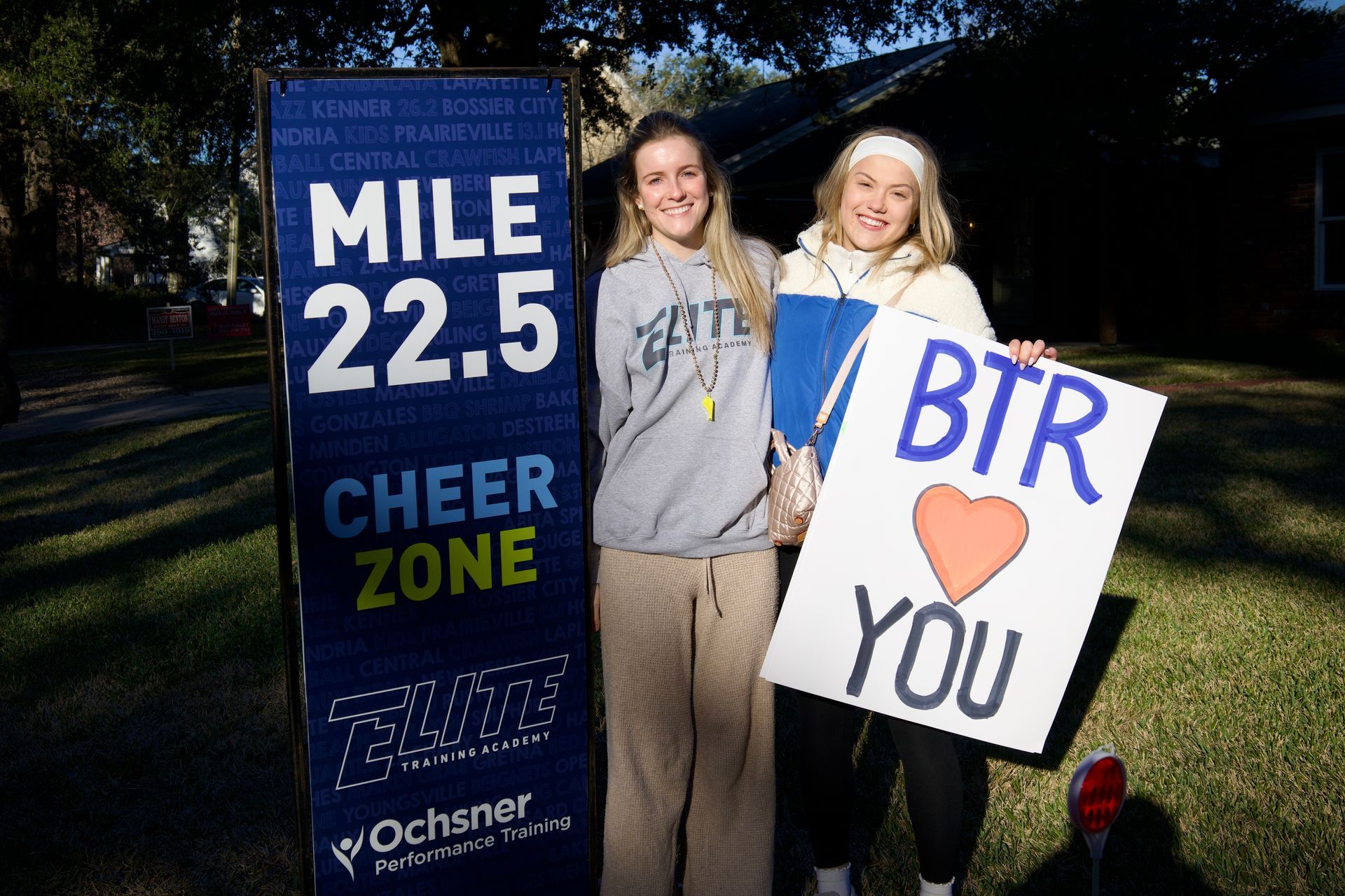 Two women at mile 22.5 cheer zone holding a “BTR ❤️ YOU” sign and standing by a race marker