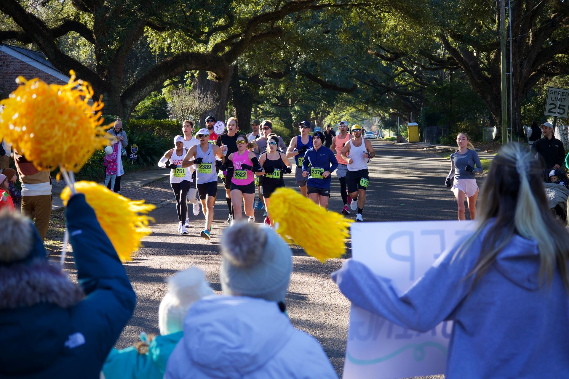 Runners in a park race toward a finish line, cheered on by spectators waving yellow pom-poms.