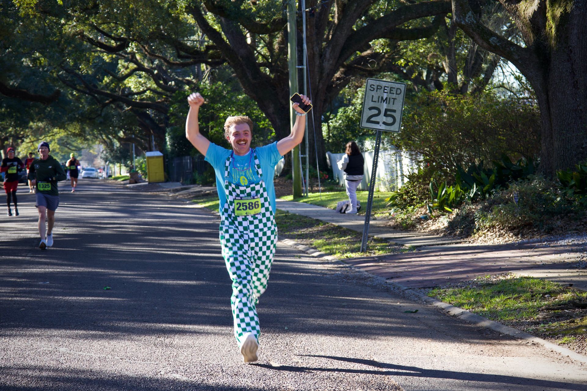 Runner in blue shirt and patterned leggings raises both arms on a tree-lined road near a 25 mph sign.